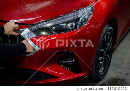 The master applies vinyl film to the headlight of a red car.  113019532