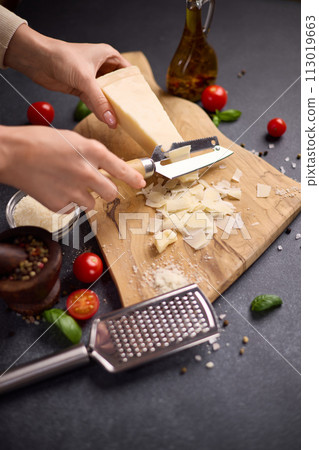 Woman slicing Piece of Traditional Italian Parmesan Hard cheese into flakes on a wooden cutting board at domestic kitchen 113019663