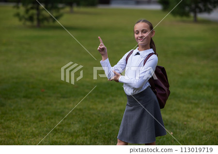 Caucasian girl in uniform and with a backpack points to an empty place outdoors after school. 113019719