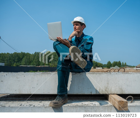 Caucasian male builder in hardhat sits on floor slabs and uses laptop at construction site. Caucasian male builder in hardhat sits on floor slabs and uses laptop at construction site. 113019723