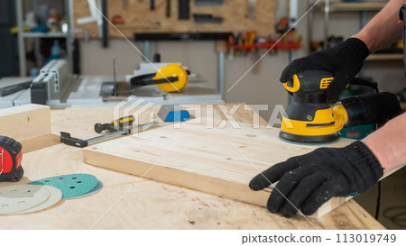 A man using an orbital wood sander in a workshop. Close-up of a carpenter's hands. A man using an orbital wood sander in a workshop. Close-up of a carpenter's hands. 113019749