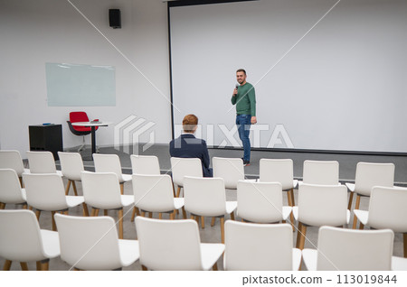 A red-haired Caucasian businesswoman sits in the front row of an empty conference room. Bearded man giving a lecture.  113019844