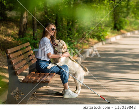 Blind young woman cuddling with guide dog while sitting on a bench. Blind young woman cuddling with guide dog while sitting on a bench. 113019848