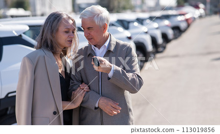 Mature Caucasian couple standing by a car outdoors. Elderly man holding car keys. Mature Caucasian couple standing by a car outdoors. Elderly man holding car keys. 113019856