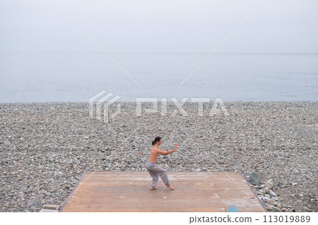 Caucasian man with naked torso practicing wushu on the seashore.  113019889