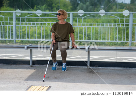 An elderly blind woman is waiting for transport at a bus stop. 113019914