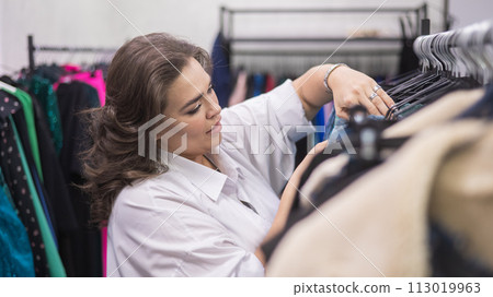 A fat woman in a plus size store chooses clothes while sorting through hangers. 113019963