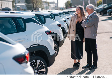 Mature Caucasian couple walks past cars outdoors, choosing a new one. Mature Caucasian couple walks past cars outdoors, choosing a new one. 113020220