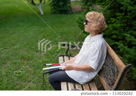 An elderly blind woman sits on a bench in the park with a folded tactile cane in her hands. 113020282