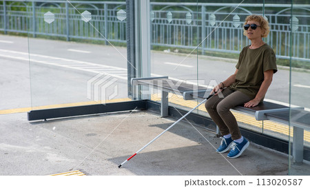 An elderly blind woman is waiting for transport at a bus stop. 113020587
