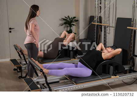 Two pregnant women are doing Pilates on a reformer. The instructor teaches prenatal yoga.  113020732