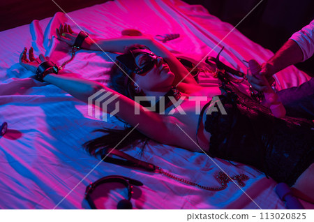 A woman in a leather mask and handcuffs lies on a bed in a blue-red light. Man holding dildo. A woman in a leather mask and handcuffs lies on a bed in a blue-red light. Man holding dildo. 113020825