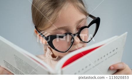 Portrait of a little girl in heart-shaped glasses reading a book. Portrait of a little girl in heart-shaped glasses reading a book. 113020853