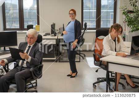 A happy pregnant woman stands in the middle of the office next to a Caucasian woman and an elderly man working at computers. A happy pregnant woman stands in the middle of the office next to a Caucasian woman and an elderly man working at computers. 113020875