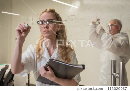 A woman chemist writes a formula on glass. An elderly Caucasian man in a protective suit is doing tests. 113020987