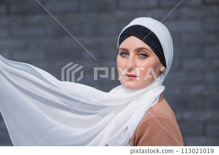 Portrait of a young blue-eyed woman in a hijab against a gray brick wall. White scarf flutters in the wind.  113021019