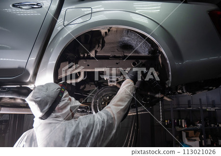 An auto mechanic applies anti-corrosion mastic to the underbody of a car. 113021026