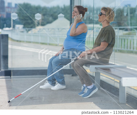 An elderly blind woman and a pregnant woman are sitting at a bus stop and waiting for the bus. An elderly blind woman and a pregnant woman are sitting at a bus stop and waiting for the bus. 113021264