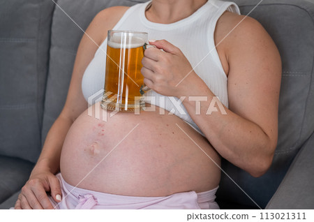 A faceless pregnant woman sits on the sofa and holds a glass of beer on her stomach. Skin rash.  113021311