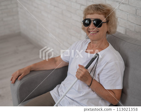 An elderly blind woman wearing sunglasses and with a tactile cane sits on the sofa.  113021358