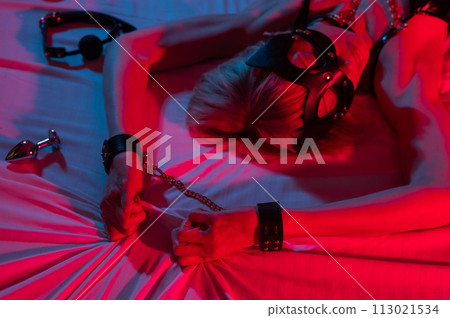 Sexy blonde woman in a leather mask and handcuffs and a collar lies in the bedroom in a red-blue neon light. Role-playing games. Sexy blonde woman in a leather mask and handcuffs and a collar lies in the bedroom in a red-blue neon light. Role-playing games. 113021534