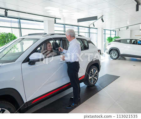 Mature Caucasian woman sits in a new car, an elderly man gives her the keys. Mature Caucasian woman sits in a new car, an elderly man gives her the keys. 113021535