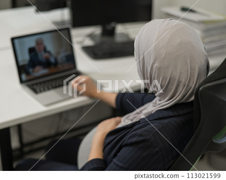A pregnant woman in a hijab communicates with a colleague via video conference on a laptop. 113021599