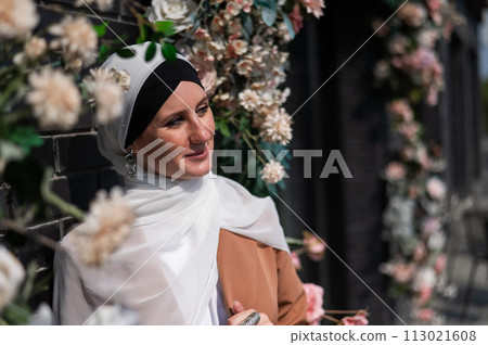 Portrait of a young caucasian woman dressed in a hijab near a wall with flowers. 113021608