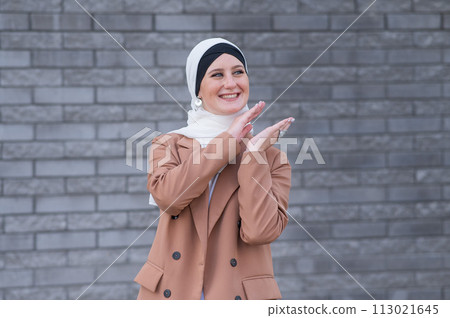 Portrait of a young blue-eyed woman in a hijab against a gray brick wall. Muslim woman happily clapping her hands. Portrait of a young blue-eyed woman in a hijab against a gray brick wall. Muslim woman happily clapping her hands. 113021645
