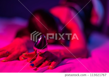 A woman in black lace lingerie lies on a bed in a red-blue neon light and holds a metal butt plug. 113021767