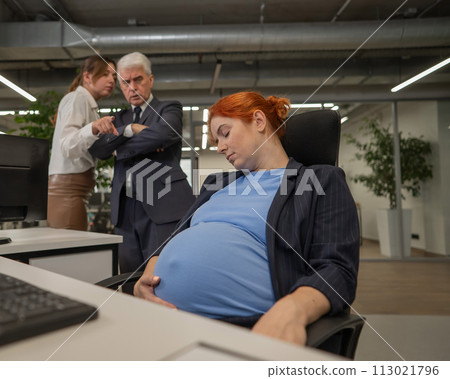 A pregnant woman sleeps at her workplace. Colleagues look disapprovingly.  113021796