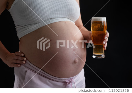 Close-up of the belly of a pregnant woman holding a glass of beer on a black background. Skin rash. Close-up of the belly of a pregnant woman holding a glass of beer on a black background. Skin rash. 113021836