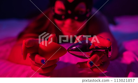 A woman lies on a bed in red and blue neon light and holds two anal plugs. BDSM concept. 113021857