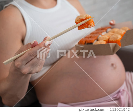 A pregnant woman sits on the sofa and eats rolls from a box. Food delivery. Close up of belly.  113021942