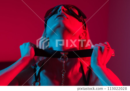 Blonde woman in leather mask putting on collar in bedroom with red blue neon light.  113022028