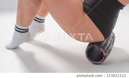 Faceless woman doing roller exercise for myofascial release on white background. Self-massage of thighs. Faceless woman doing roller exercise for myofascial release on white background. Self-massage of thighs. 113022122