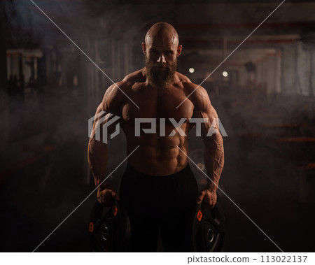 Muscular bald shirtless man holding weight plates. A bodybuilder shows off his form in the gym. 113022137