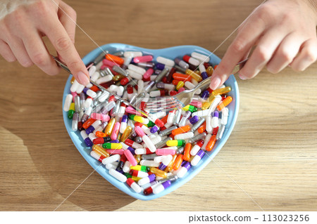 Female hands holding table knife and fork above heart shaped bowl with pile of medicine. Female hands holding table knife and fork above heart shaped bowl with pile of medicine. 113023256