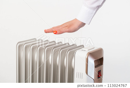 A woman's hand with a manicure warms itself against the background of an electric heater, white background. The concept of heating in apartments during the autumn and winter season, industry A woman's hand with a manicure warms itself against the background of an electric heater, white background. The concept of heating in apartments during the autumn and winter season, industry 113024767