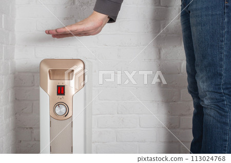 A man warms his hands on an electric oil heater at home. The concept of cold in apartments in autumn and winter. Turn on the heating. Copy space for text 113024768