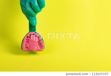 The hand of a dentist doctor in a green medical glove holds a dental spoon with an alginate plastic mass for making a plaster cast of the patient s jaw on a yellow background. Concept of malocclusion, The hand of a dentist doctor in a green medical glove holds a dental spoon with an alginate plastic mass for making a plaster cast of the patient s jaw on a yellow background. Concept of malocclusion, 113025337