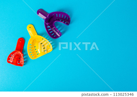 Multi-colored dental spoons for taking an impression of the dental jaw on a blue background. Orthodontics in dentistry. Dental prosthetics and crown manufacturing, close-up. Copy space for text Multi-colored dental spoons for taking an impression of the dental jaw on a blue background. Orthodontics in dentistry. Dental prosthetics and crown manufacturing, close-up. Copy space for text 113025346