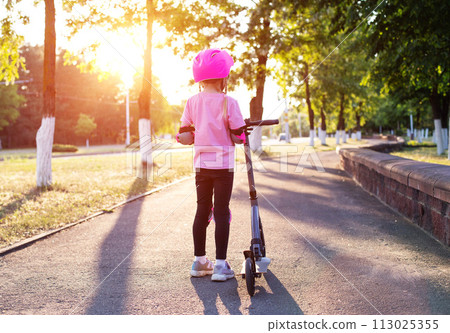 A little girl in a protective pink outfit and a helmet stands with her back with a scooter on a sunny alley. A little girl in a protective pink outfit and a helmet stands with her back with a scooter on a sunny alley. 113025355