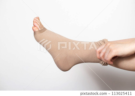 A woman puts on compression stockings for legs with varicose veins. Pain and swelling in the legs, phlebology. White background, close-up. Bloodstream 113025411