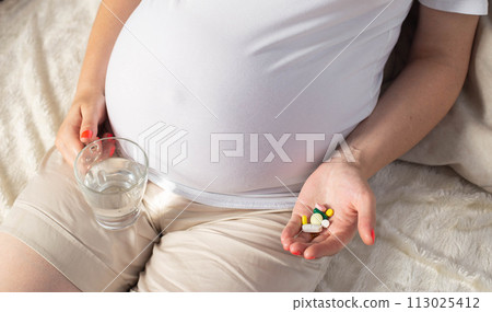 A pregnant girl holds pills in her palm and another glass of water. Concept of taking vitamins, iron and folic acid during pregnancy, close-up A pregnant girl holds pills in her palm and another glass of water. Concept of taking vitamins, iron and folic acid during pregnancy, close-up 113025412