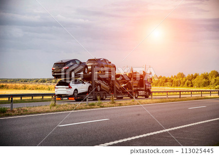 An auto transporter semi-trailer truck transports broken damaged cars from Europe in the summer against a sunset background. Car import and export concept, shipping 113025445