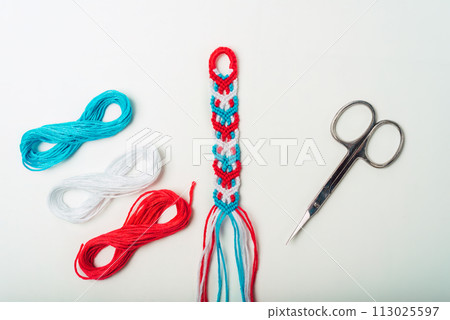 Unfinished woven DIY friendship bracelet with boho pattern next to scissors on white background 113025597