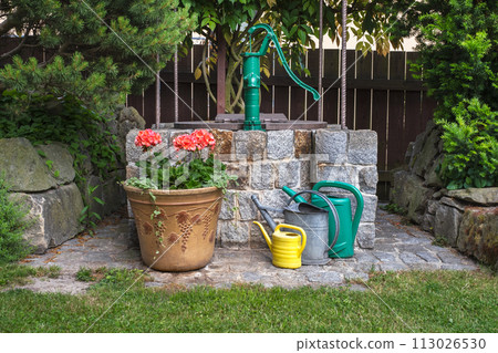 Stone well in a summer garden with potted flowers and watering cans Stone well in a summer garden with potted flowers and watering cans 113026530