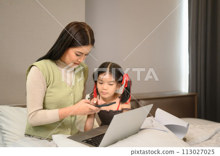 Young mother working from home and using laptop with cute little daughter in bedroom Young mother working from home and using laptop with cute little daughter in bedroom 113027025