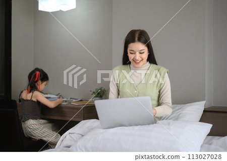 Beautiful mother working on laptop and her preschooler daughter doing homework in bedroom 113027028